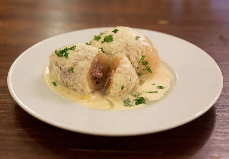 Cepelinai in sour-cream sauce, one cut to show the meat filling; elongated potato-dough shape. Cooked.