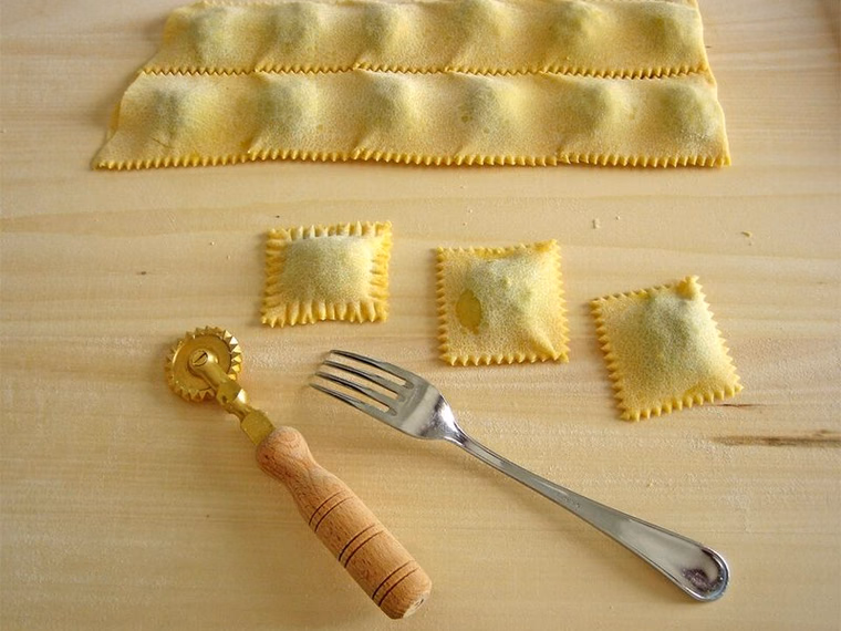 Uncooked square ravioli cut from a filled pasta sheet, with a fluted cutter and fork. Shape and crimped edge very clear.